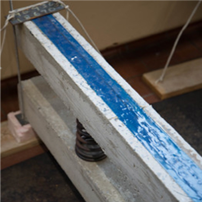 Photo of one long, narrow white concrete bar stacked on top of another, with a thick spring in between. The top bar is filled with a stripe of blue material down the middle.